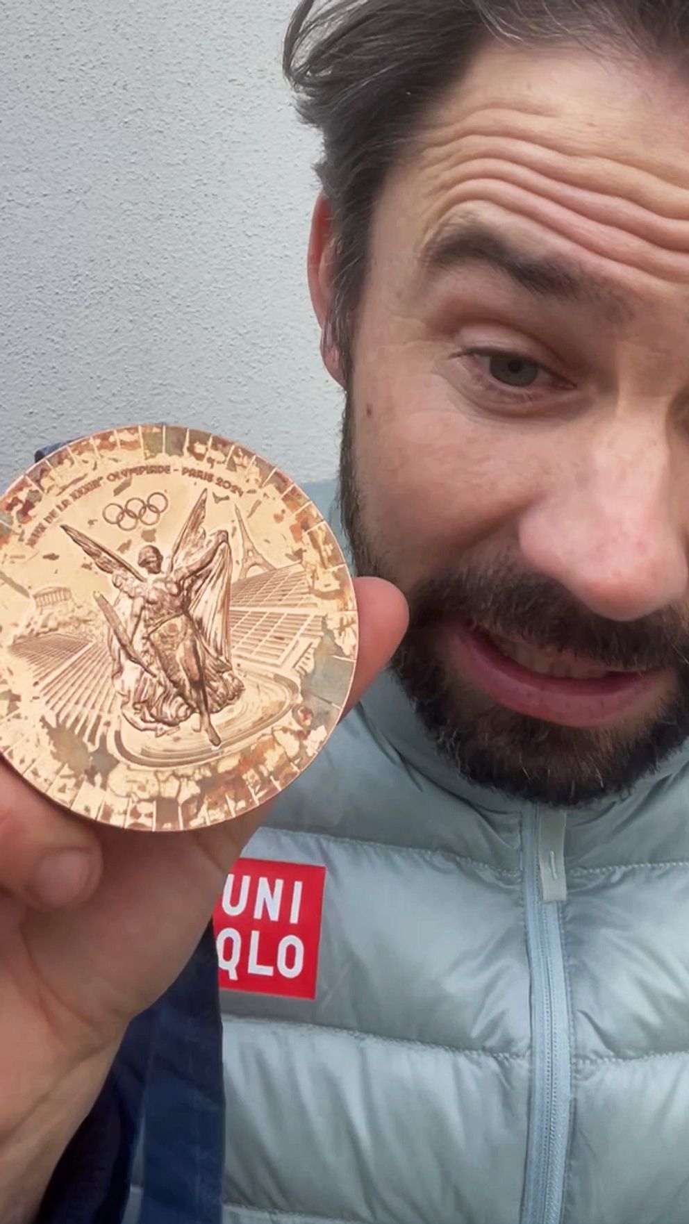 En person håller upp en bronsmedalj med en symbol av en örn, som representerar en prestation eller vinst. Personen står framför en neutral bakgrund iförd en lätt jacka.