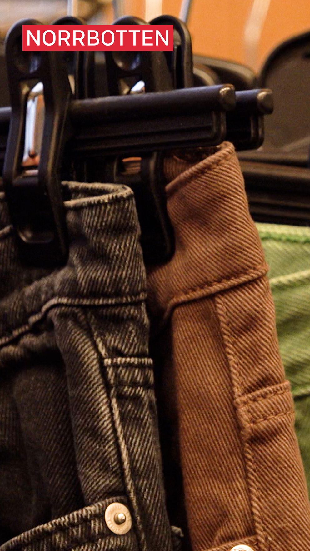 En närbild av två par hängande byxor, ett par i mörk denim och ett par i brunt tyg, hållna av klädklämmor. Ovanför byxorna finns en röd text som säger ”NORRBOTTEN”.