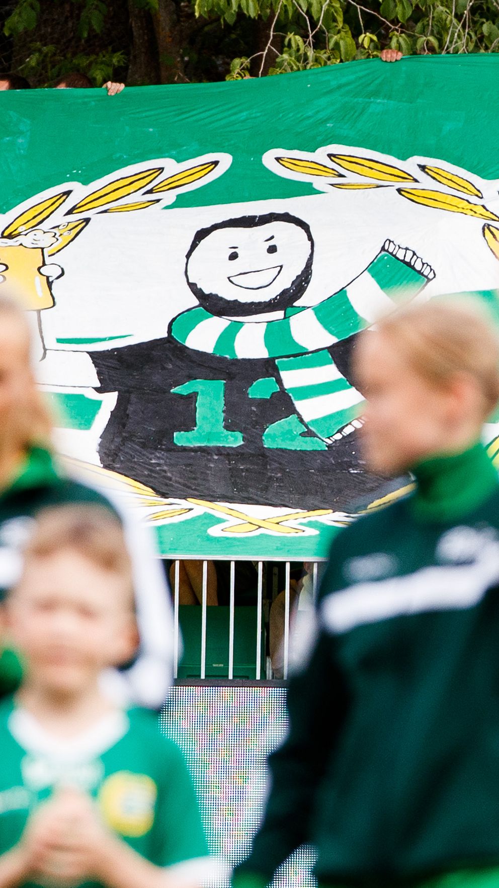 En grup av unga fotbollsspelare i gröna och svarta kläder intar planen framför en stor banner. Bannern visar en tecknad figur med en tröja med nummer 12, omgiven av en krans av lagerblad. Figuren jublar och håller upp en pokal, vilket indikerar firande och stolthet.