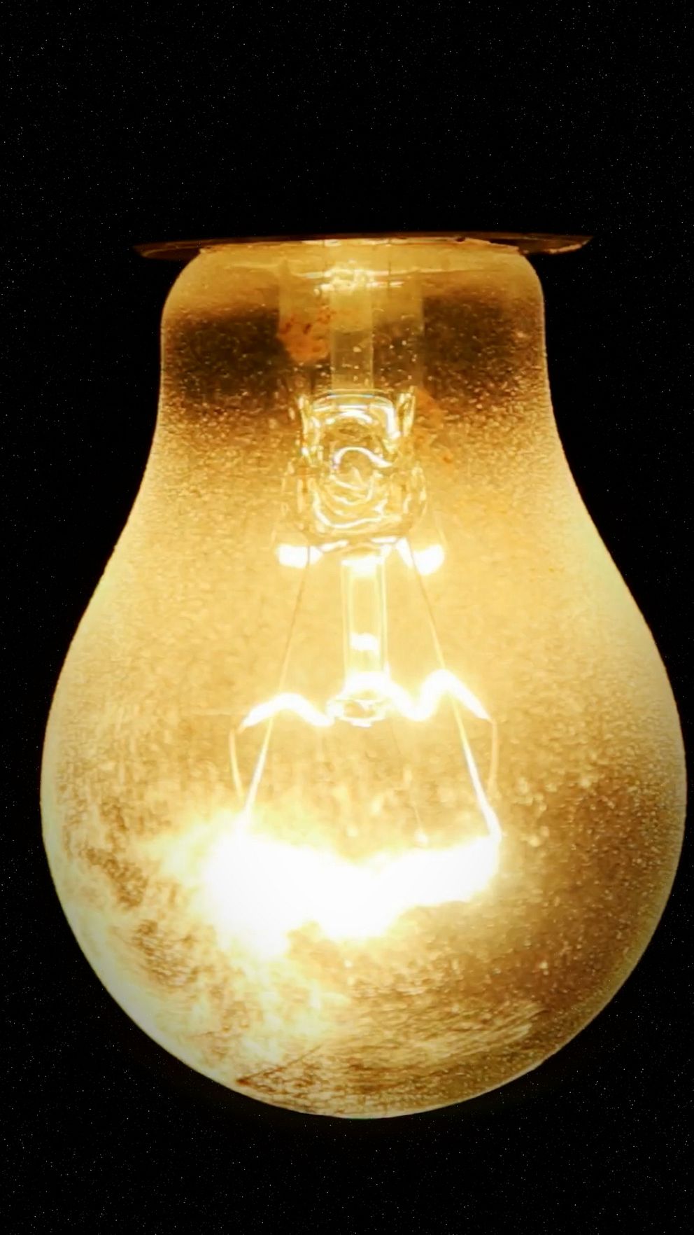Glödlampa som lyser med ett varmt, gyllene sken mot en svart bakgrund. Lampan har en lätt dimmig yta och visar den synliga glödtråden inuti.