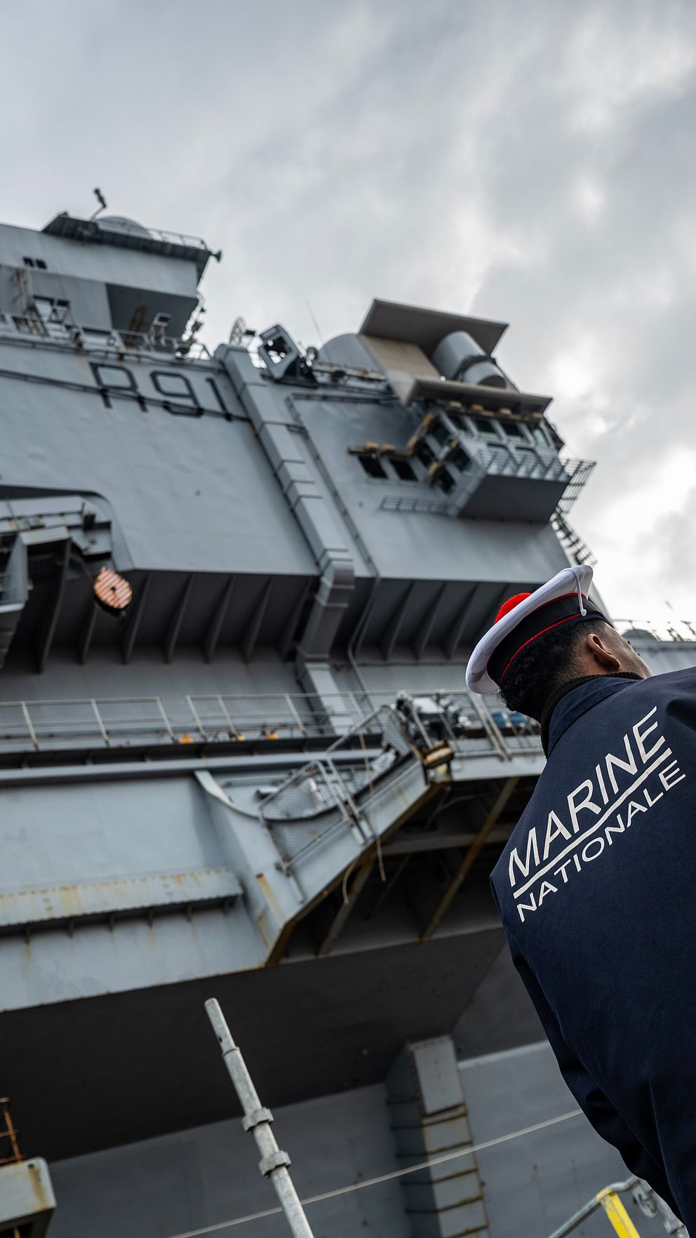 En person i uniform står vid ett stort militärfartyg under molnig himmel.
