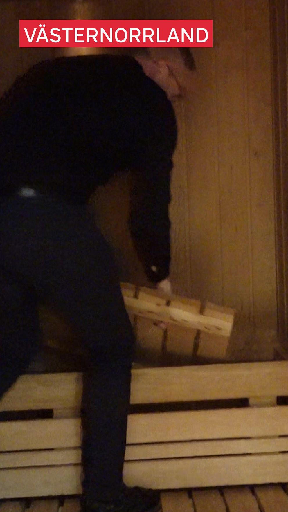 En person står i en bastu och lyfter en träbit från en stapel av träplankor. Texten ”VÄSTERNORRLAND” är placerad högst upp i bilden.