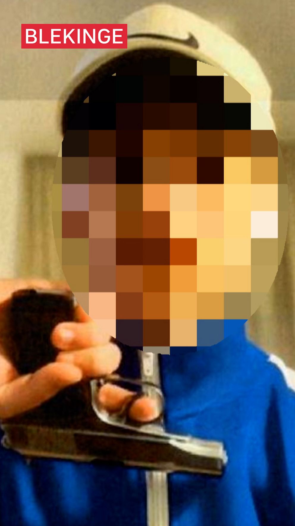 En pixlad bild visar en individ i en blå jacka och vit keps som håller upp ett pistolliknande föremål. I det övre vänstra hörnet finns en etikett med texten ”Blekinge”.