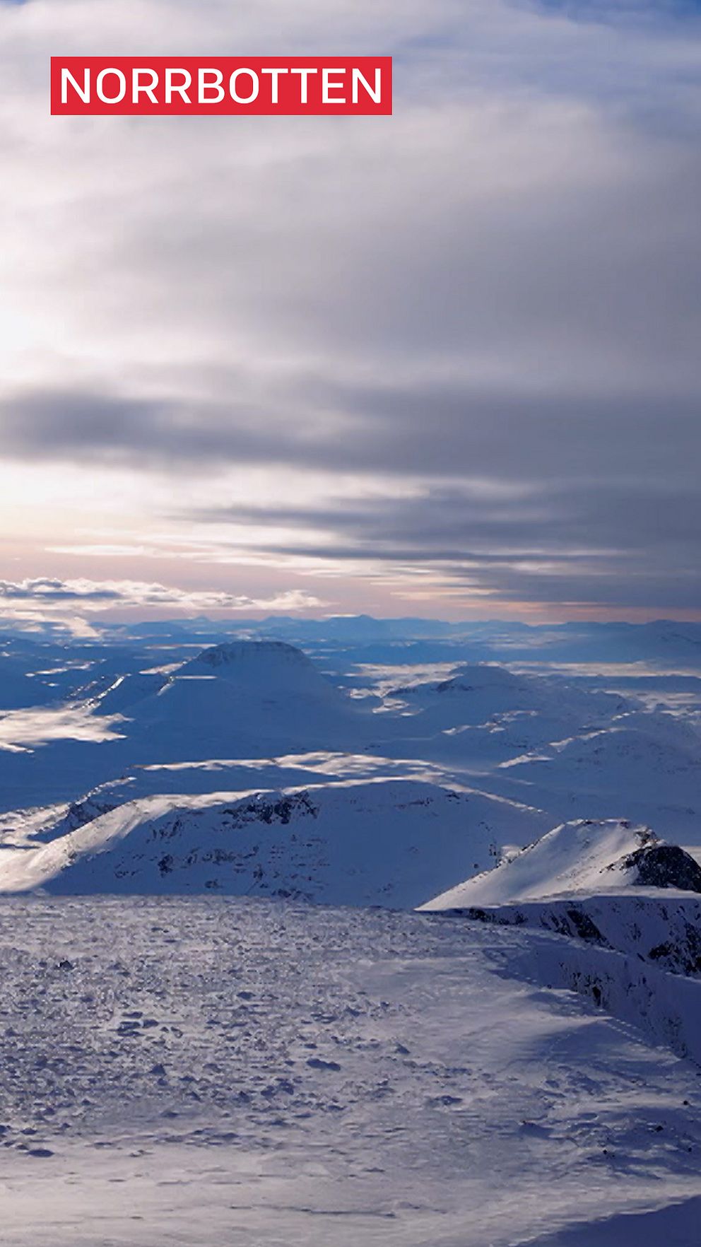 Snöklädda fjäll och bergslandskap i Norrbotten under en mulen himmel, med ett mjukt ljus från den delvis gömda solen.