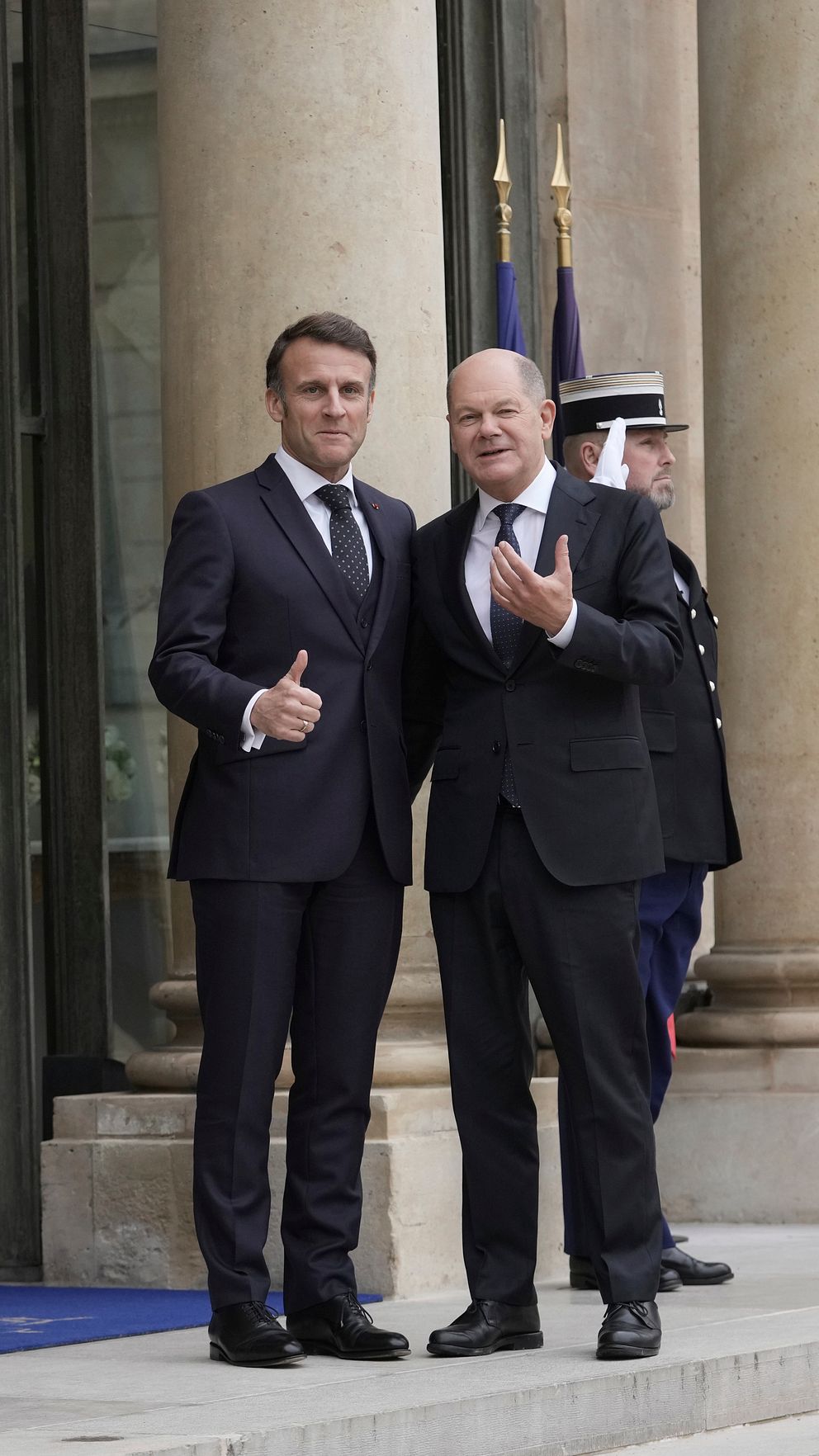 Emmanuel Macron och Olaf Scholz poserar tillsammans framför en byggnad, båda ger tummen upp, omgivna av vakter i ceremoniella uniformer.