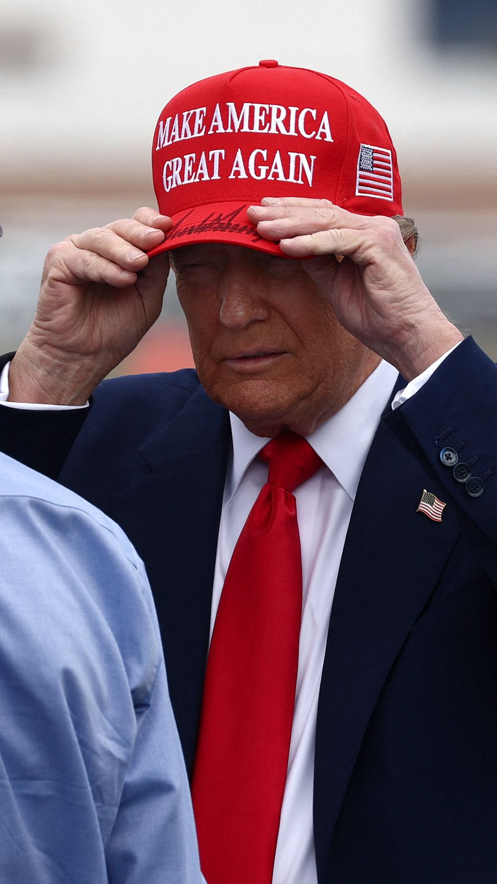 Två personer står utomhus och samtalar. Den ena har på sig en keps med siffran ”45” tryckt på baksidan. Den andra bär en röd keps med texten ”Make America Great Again” och en kostym med röd slips. De befinner sig på en plats som verkar vara en flygplats eller liknande.