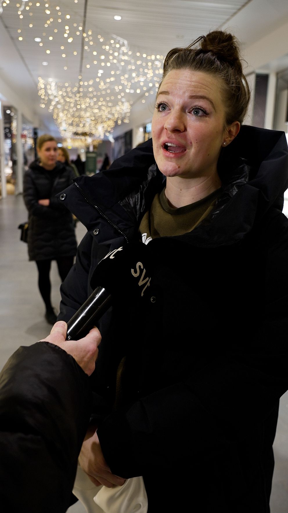 En person står i en upplyst gång med juldekorationer i bakgrunden. Personen bär en stor svart jacka och blir intervjuad av en journalist som håller en mikrofon. I bakgrunden syns människor som går förbi.