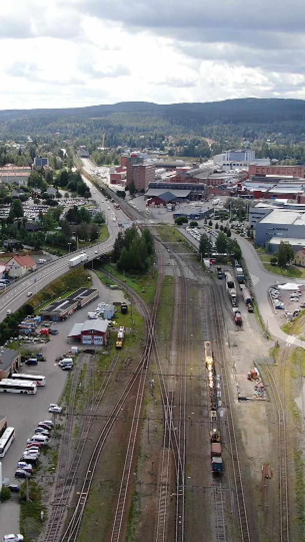 En vy över en stad, Ludvika, med bostadsområden, affärsbyggnader och en järnväg. I förgrunden syns byggnader och parkeringsområden, medan bakgrunden visar fabriksbyggnader och gröna kullar. Järnvägen sträcker sig genom bilden med tåg som är synliga i rälsen.