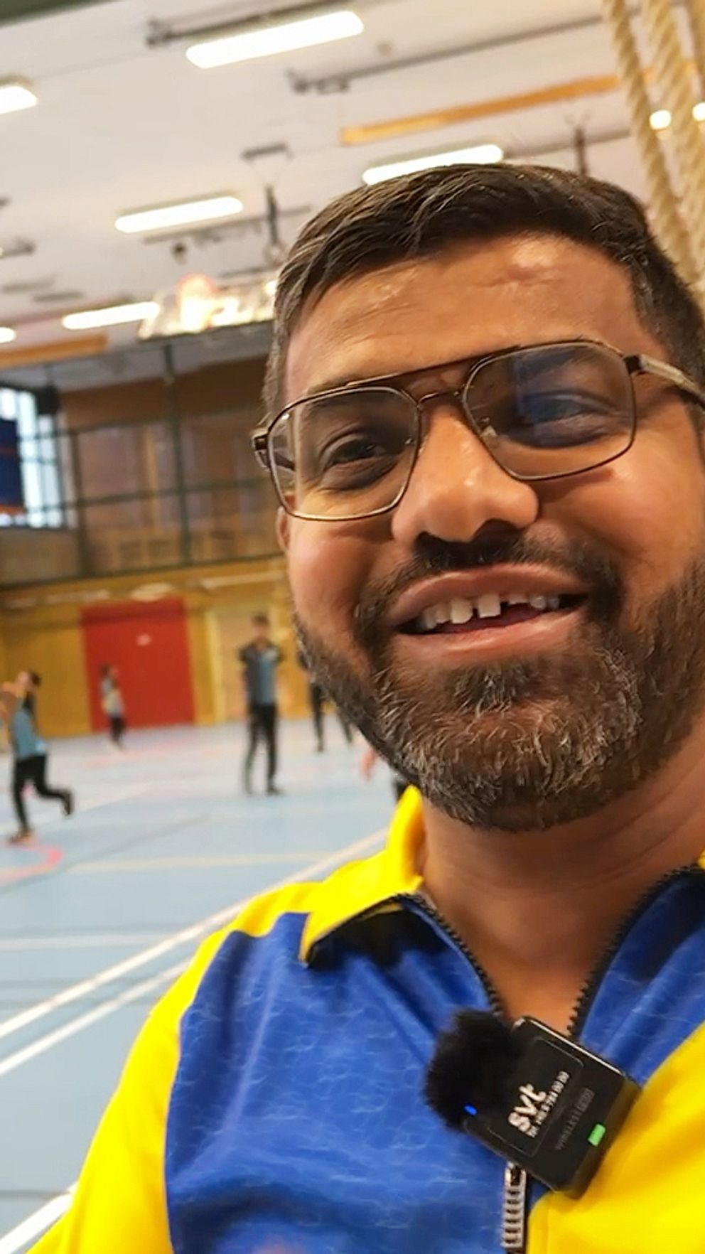 Cricketledaren Syed Salami i en gul och blå cricketträningsdräkt i en sporthall. I bakgrunden syns en grupp människor som tränar cricket på en inomhusplan med linjer och mål.