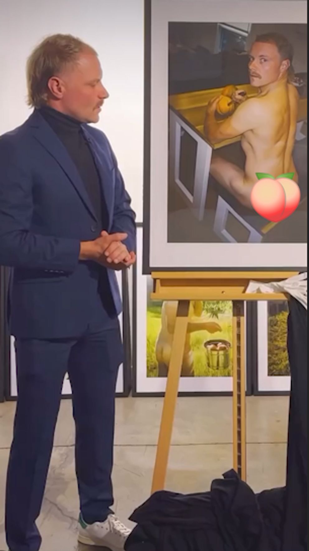 En man i kostym står i ett galleri och betraktar ett inramat fotografi på ett staffli. Fotot föreställer en muskulös person sittande vid ett bord, delvis täckt med en emoji. I bakgrunden syns fler inramade bilder på golvet.