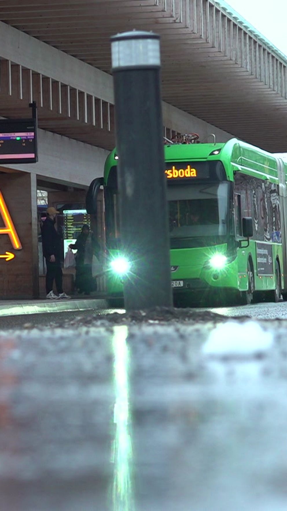 En buss vid en hållplats med gröna detaljer, där passagerare väntar på att stiga på. I bakgrunden syns digitala tidtabeller, och stora bokstäver som formar ordet ”E A” i orange neonskrift. Vattnet på marken reflekterar den kalla atmosfären.