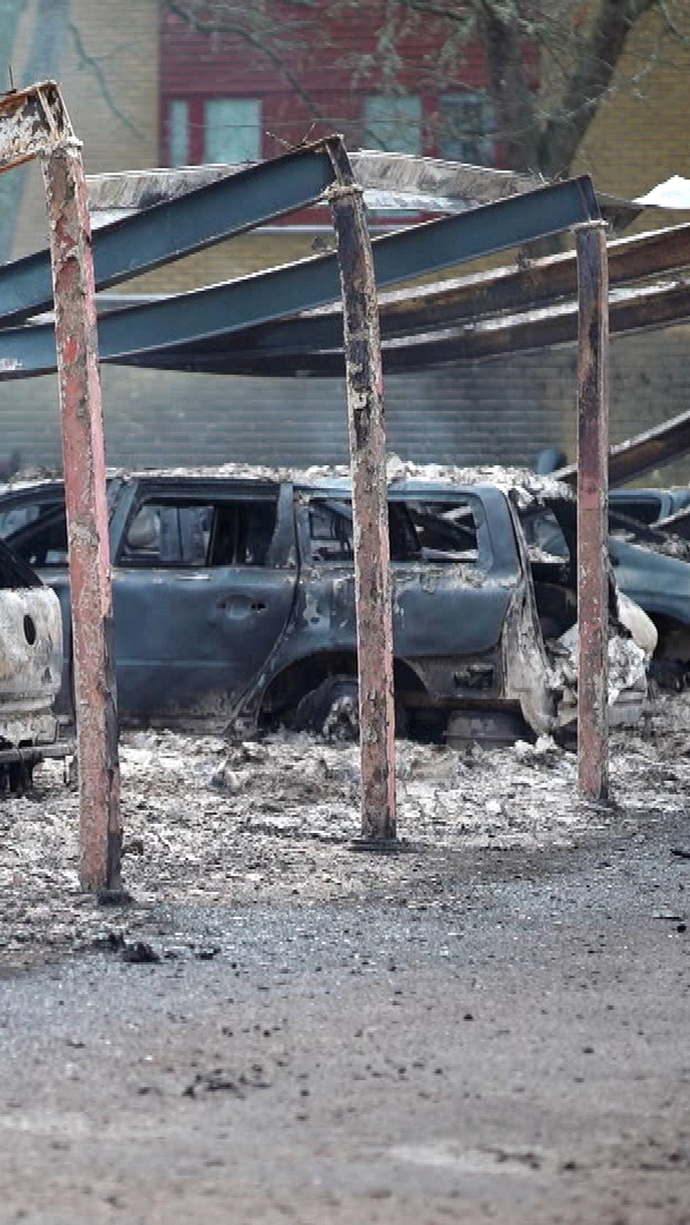 Rader av brända bilar står under en delvis kollapsad carport, omgivna av aska och rök efter en brand. Kvarstående ramar av carporten är synliga i förgrunden, medan de skadade bilarna ligger övergivna. Bakgrunden visar otydliga byggnader som tyder på att området har drabbats av förödelse.