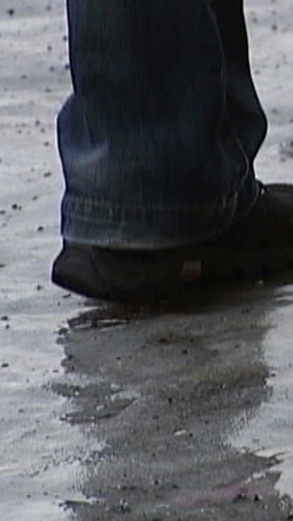 Fötter i jeans och skor som går över en blöt yta, reflektioner syns på den regnvåta marken.