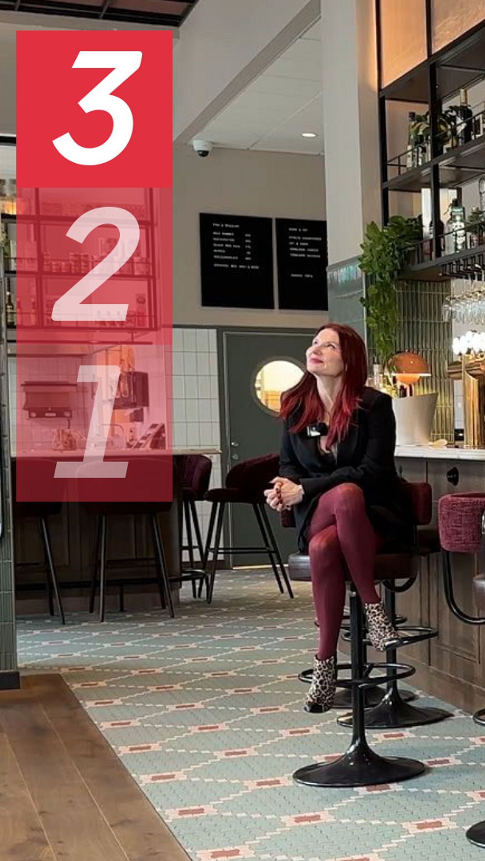 En stilren bar med en lång bänkdisk, inredd med trä och metall. Hyllor med alkoholflaskor syns bakom disken, där glas hänger i rader. Emma Engdahl, kärleksexpert, sitter på en barstol klädd i svarta kläder, vänd mot kameran. Golvet är dekorerat med ett mönstrat kakel och det finns växter i lokalen som bidrar till den moderna atmosfären.