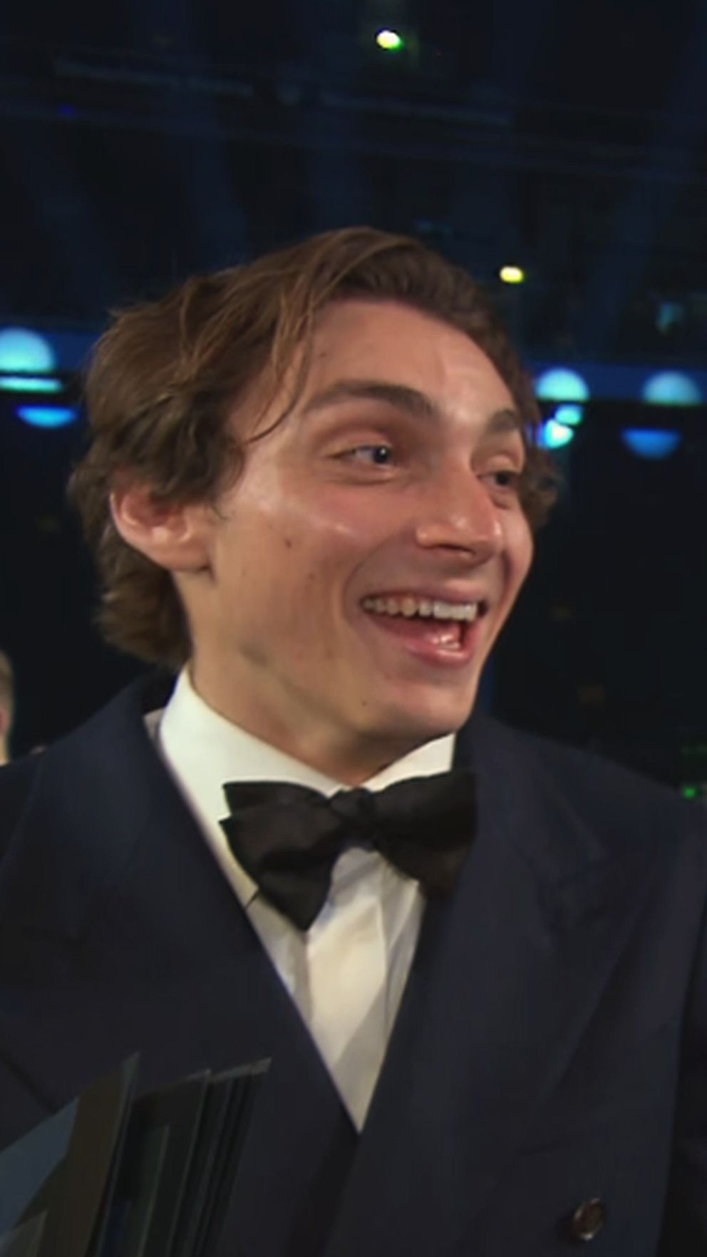 En man i smoking och en kvinna i en svart klänning intervjuas vid en gala, med fokus på kvinnan som håller en mikrofon. I bakgrunden syns en festlig publik och festlig belysning.
