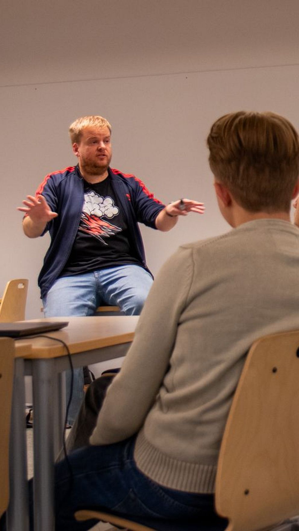 Läraren Gustav Wigren står vid en whiteboard och gestikulerar medan han pratar med en grupp studenter som sitter vid bordet. De lyssnar uppmärksamt i ett klassrum