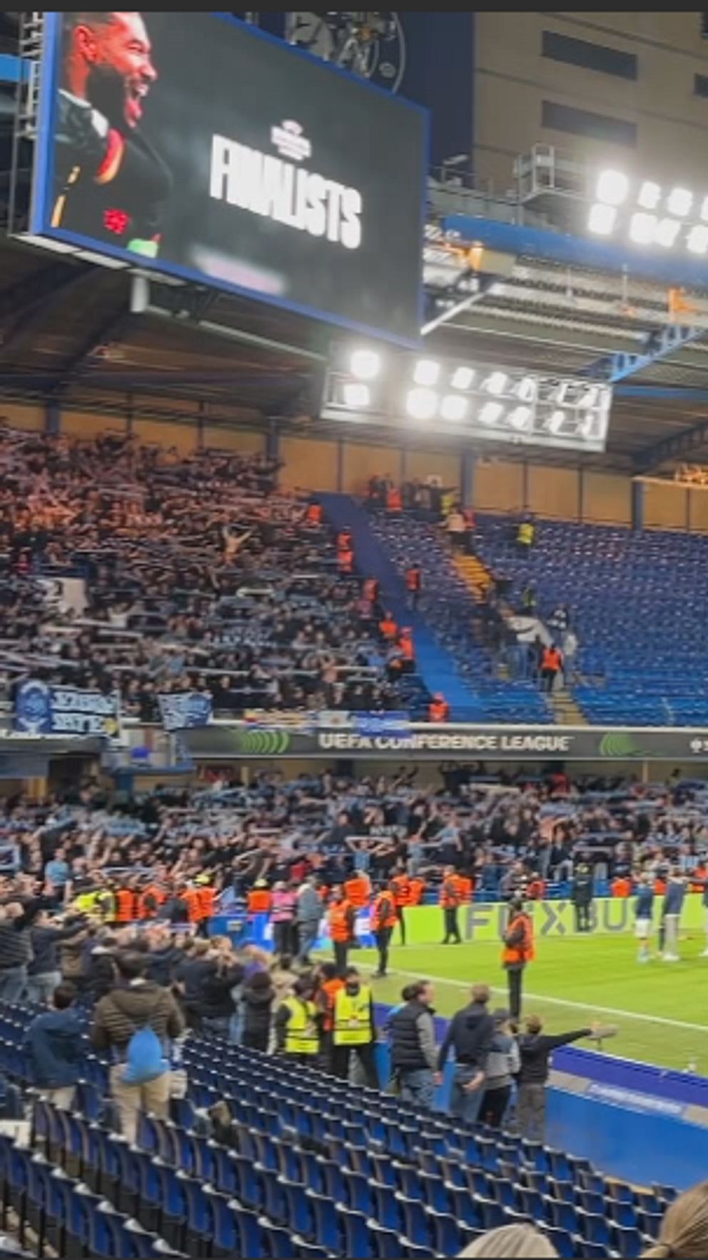 Fotbollsarena med spelare och funktionärer på planen, delvis fyllda läktare kvällstid.