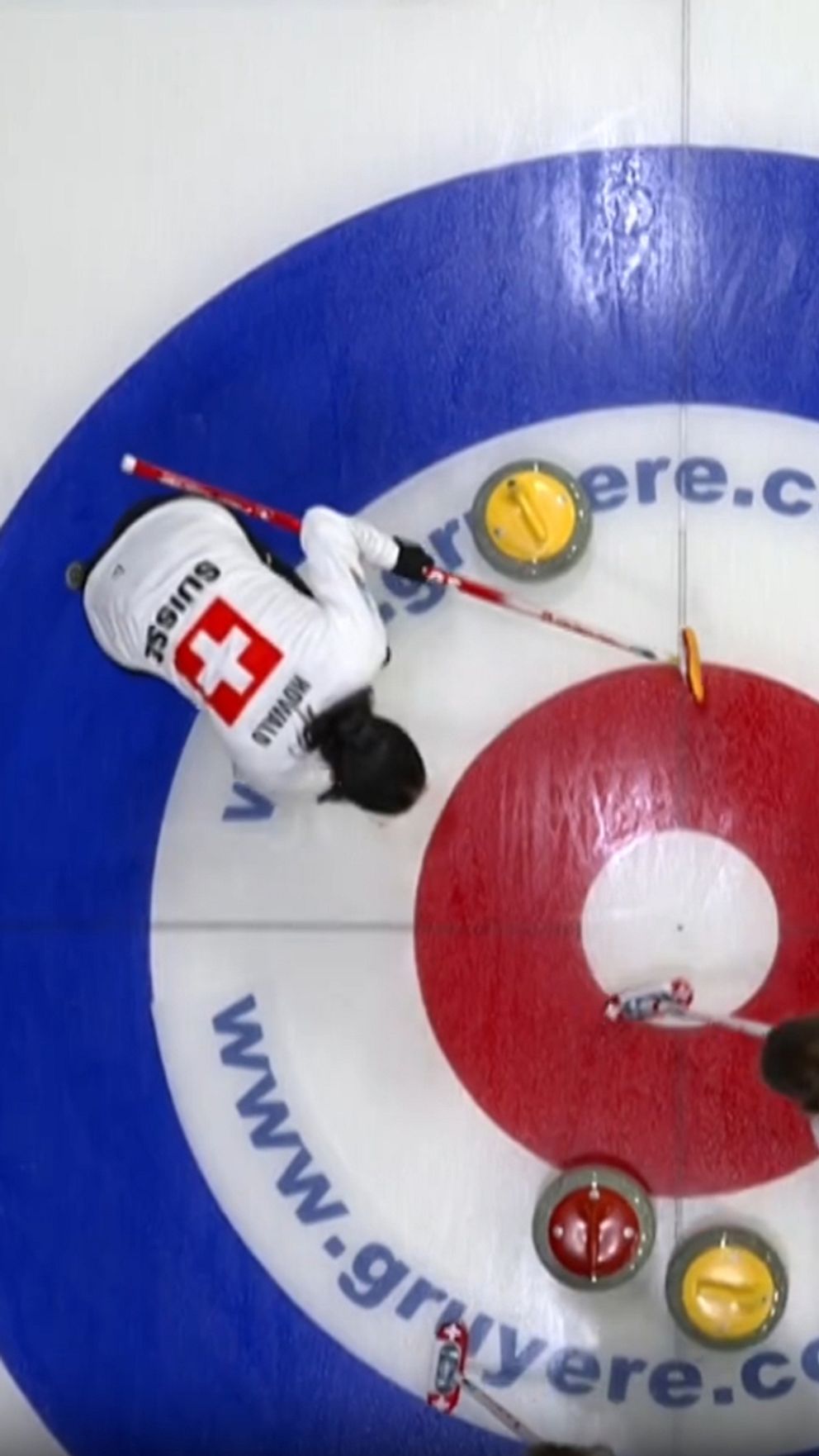 En delad bild som visar en curlingmatch. Till vänster syns två spelare i vit utrustning från Schweiz som positionerar sina stenar på isen i en curlingring med blå och röd cirkel. Det finns en gul och en röd sten nära mitten av cirkeln. Till höger ses ett lag i mörka kläder som står vid sin bänk och följer spelet, med fokus på en tavla där poäng och laginformation visas.