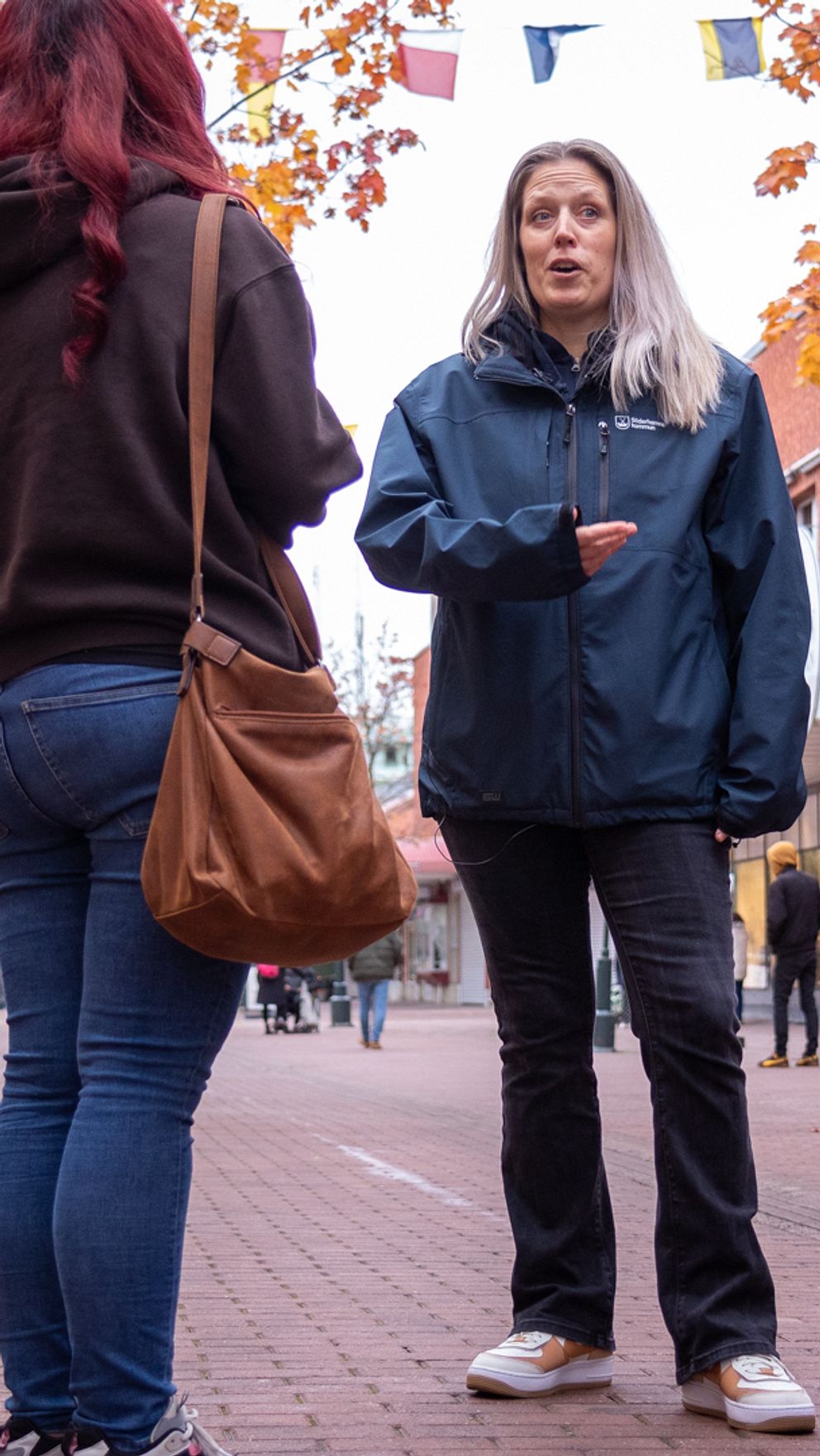 Tre personer står på en gata omgiven av höstlöv och pratar med varandra. Till höger ligger en svart vape enhet lutad mot ett golv.