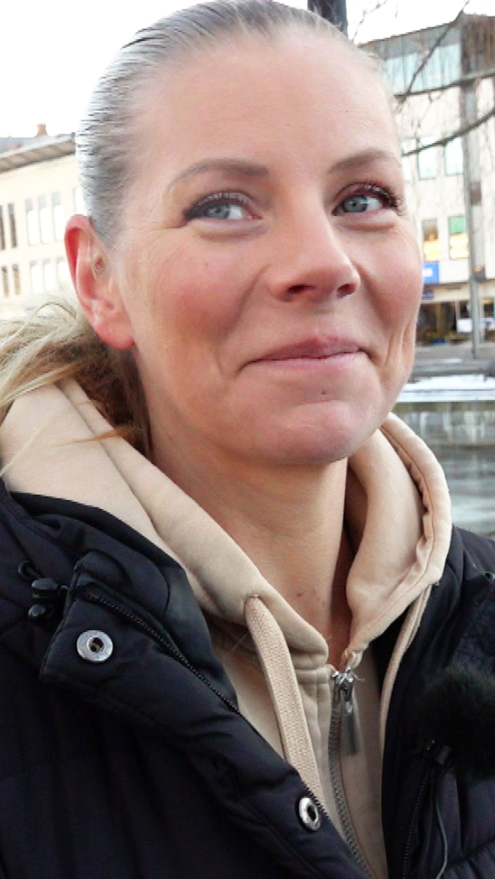 En kvinna står i förgrunden vid en vattenyta som reflekterar en byggnad i bakgrunden. Vattnet har ringar som skapas av en utter vid ytan, och snö ligger längs kanten. I bakgrunden syns fler byggnader i en stadsmiljö.