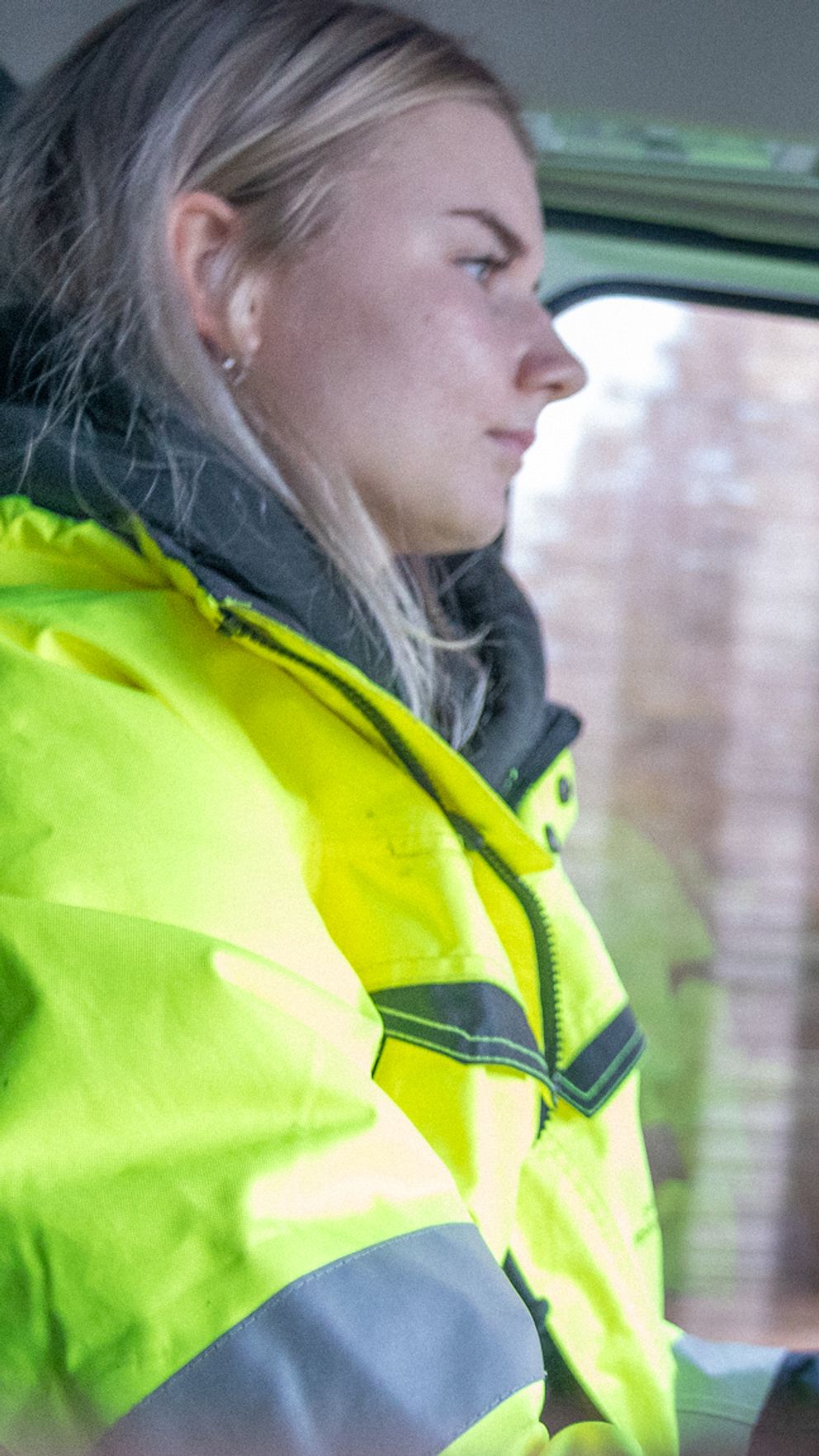 En person i en gul säkerhetsjacka sitter bakom ratten i en lastbil, med fokus på körfältet utanför. Bakgrunden visar suddiga träd som passerar, vilket antyder att fordonet är i rörelse.