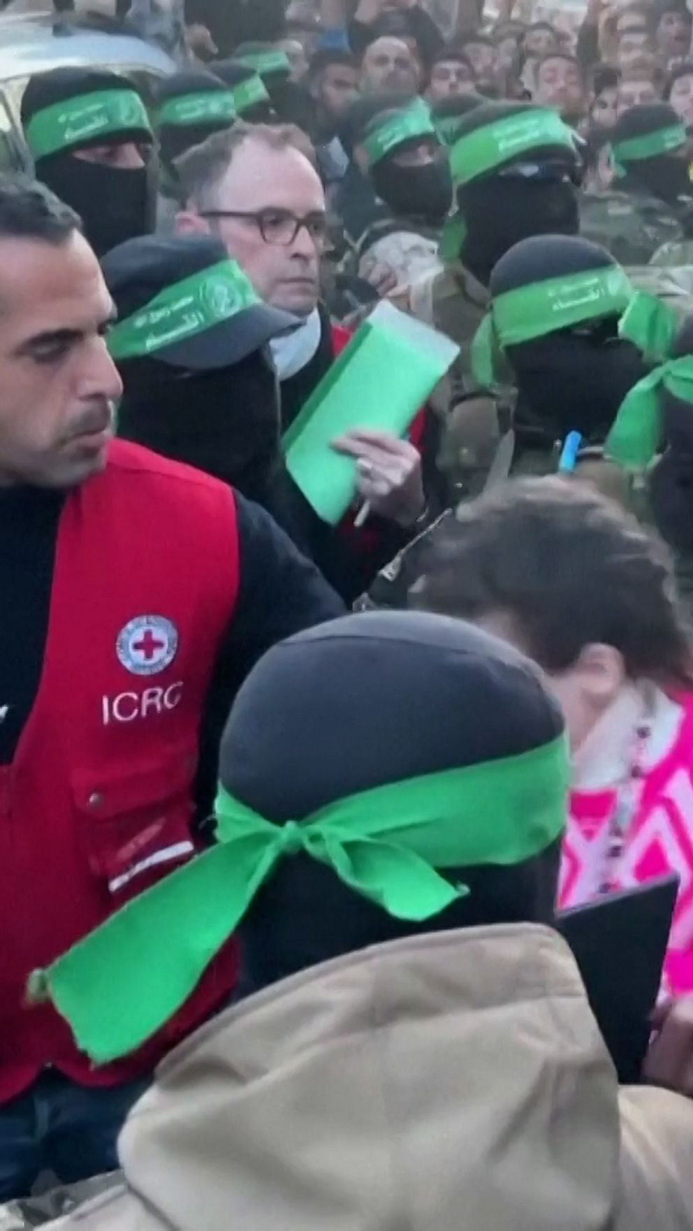 En stor grupp personer med svarta masker och gröna band runt huvudet omger och interagerar med en kvinna i en rosa tröja nära en bil. En man i röd väst från Röda Korset är synlig i närheten. Bakgrunden består av en folksamling.