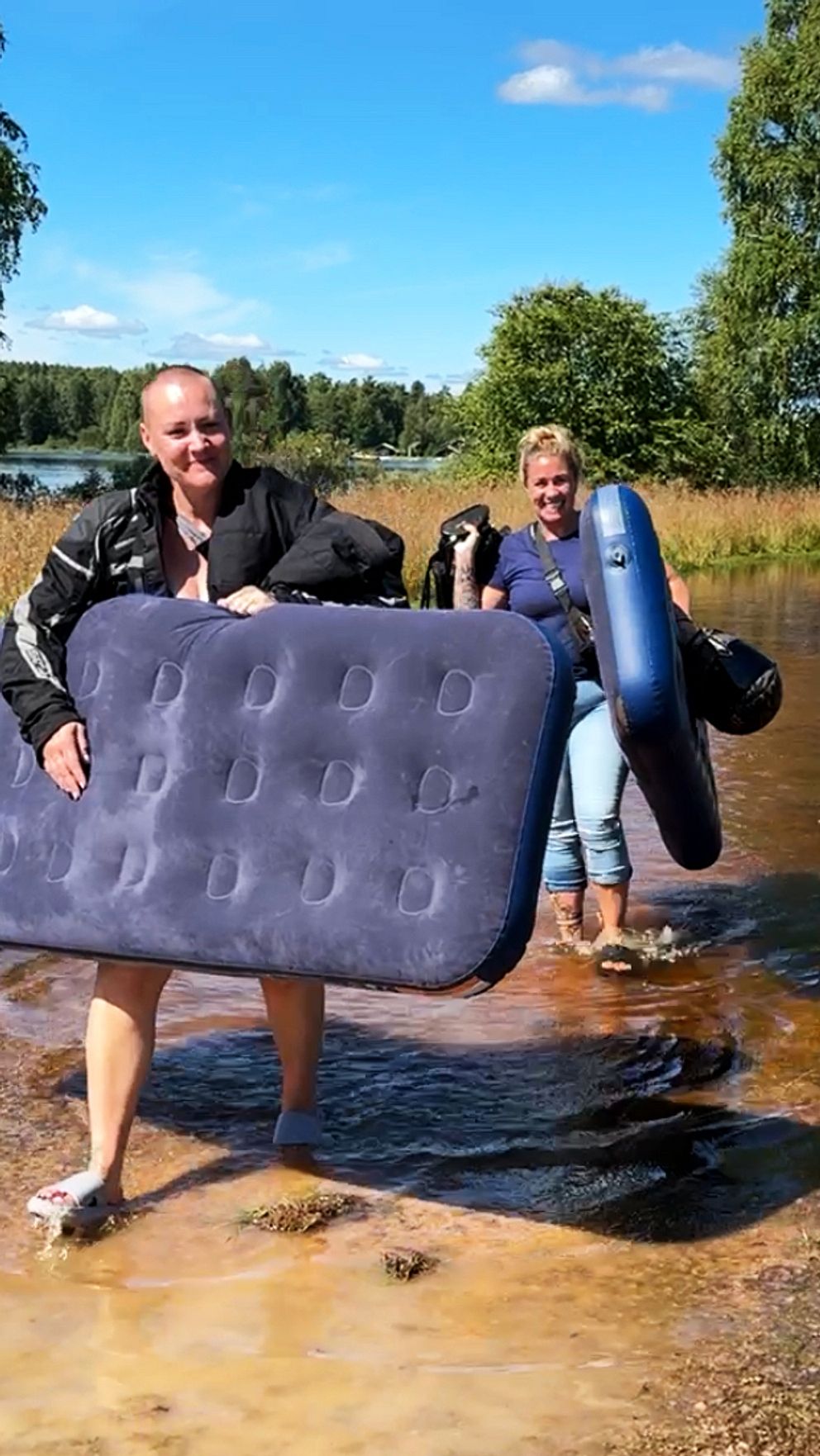 Två campinggäster vid Dansbandsveckan i Malung bär på sina luftmadrasser samtidigt som de vadar genom vattnet som översvämmat campingen.