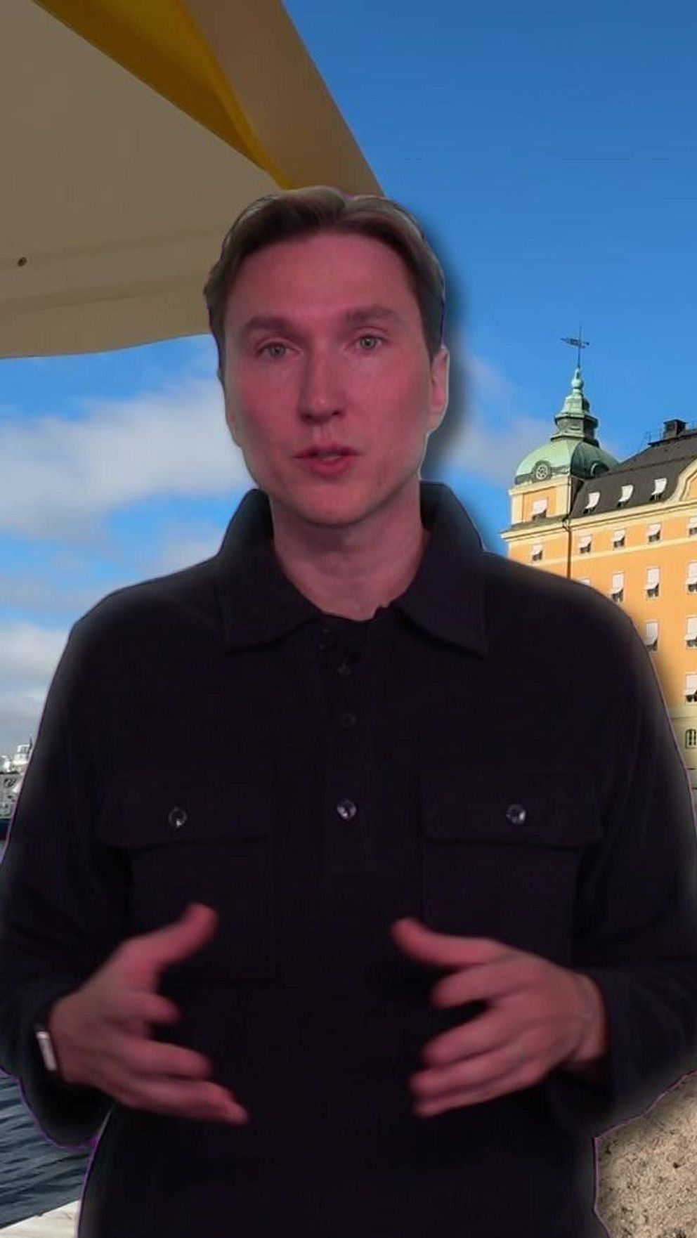 En person står framför en vattenspegel med en röd och gul byggnad i bakgrunden. Byggnaden har flera spira-toppar och en grön taklinje, omgiven av träd. I förgrunden syns en solstol och en skuggig yta under ett tak. Det är klart väder med blå himmel.