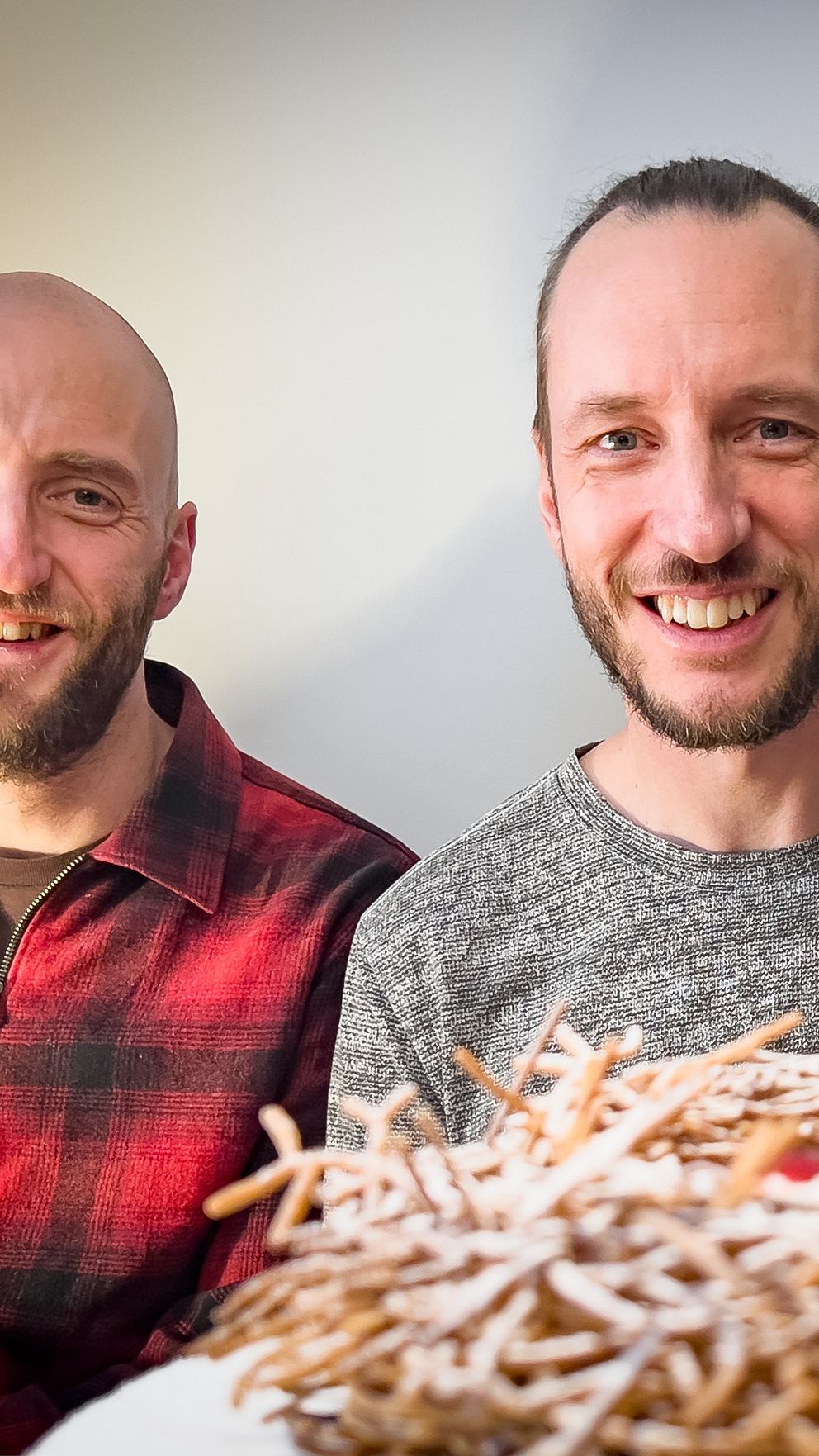 Bilden visar två personer som sitter vid ett bord, med en detaljerad dekoration av fåglar i pepparkaka framför dem. Dekorationen är placerad på ett vitt fat inkluderar en stor fågel och flera mindre fåglar i en näste-liknande uppställning. Bakgrunden är ljus och enkel, vilket framhäver konstverket.