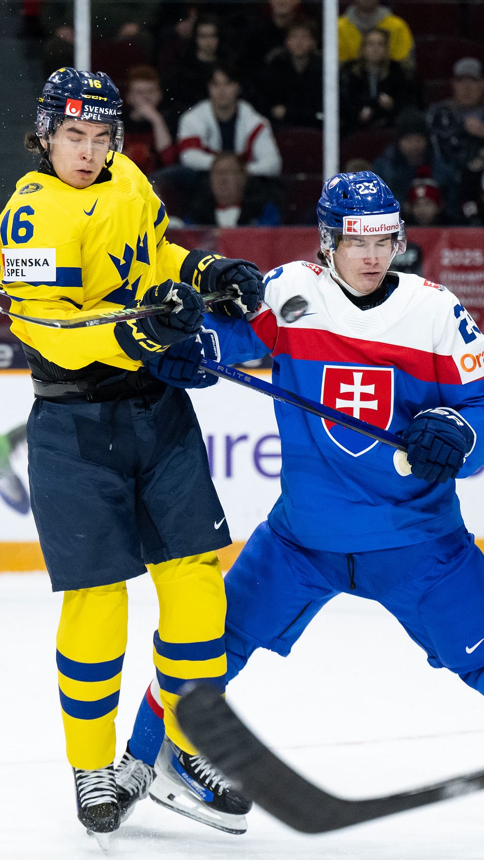 Spelare i blå och gula hockeyutrustningar kämpar om pucken under en match. Den svenska spelaren i gult står i en defensiv position medan den slovakiska spelaren i blått är aktiv i en tackling. I bakgrunden syns en publik på läktaren, vilket ger en indikation på den intensiva atmosfären i arenan.