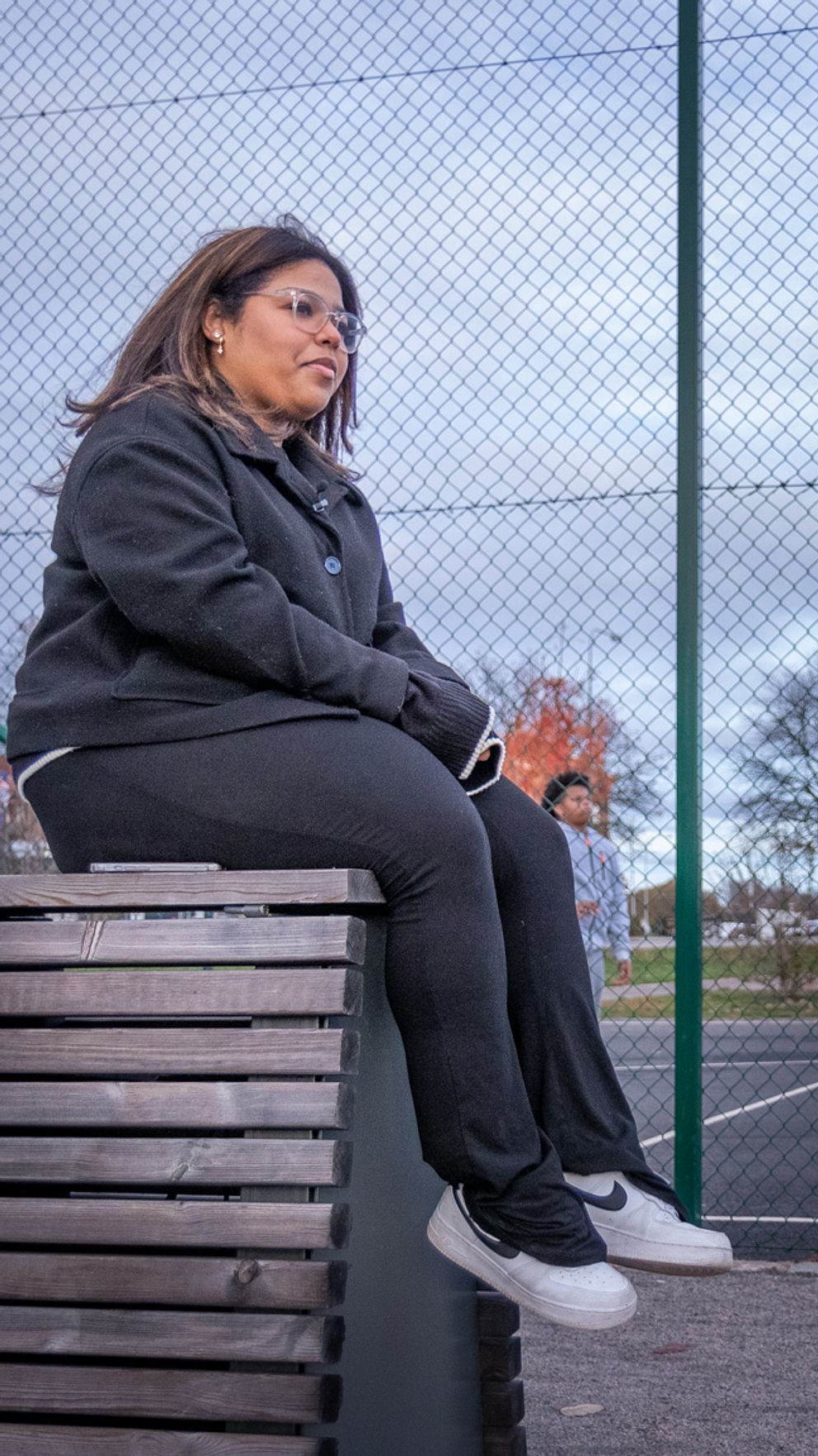 En person sitter på en bänk i förgrunden, medan två andra personer spelar basket i bakgrunden. En av basketspelarna kastar bollen mot korgen, medan den andra står redo att ta emot den. Planen är omgiven av nät, och omgivningen har träd och byggnader i bakgrunden.