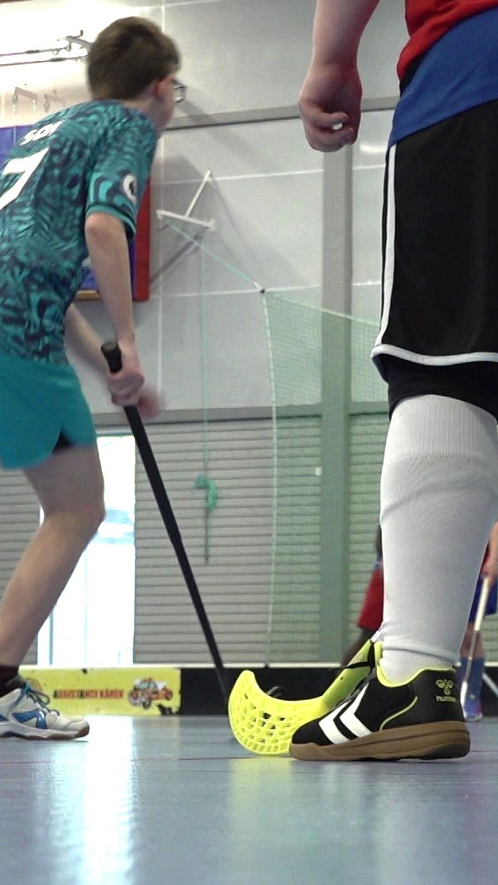 Unga spelare spelar innebandy i en idrottshall. Klubbfäktning nära målet.