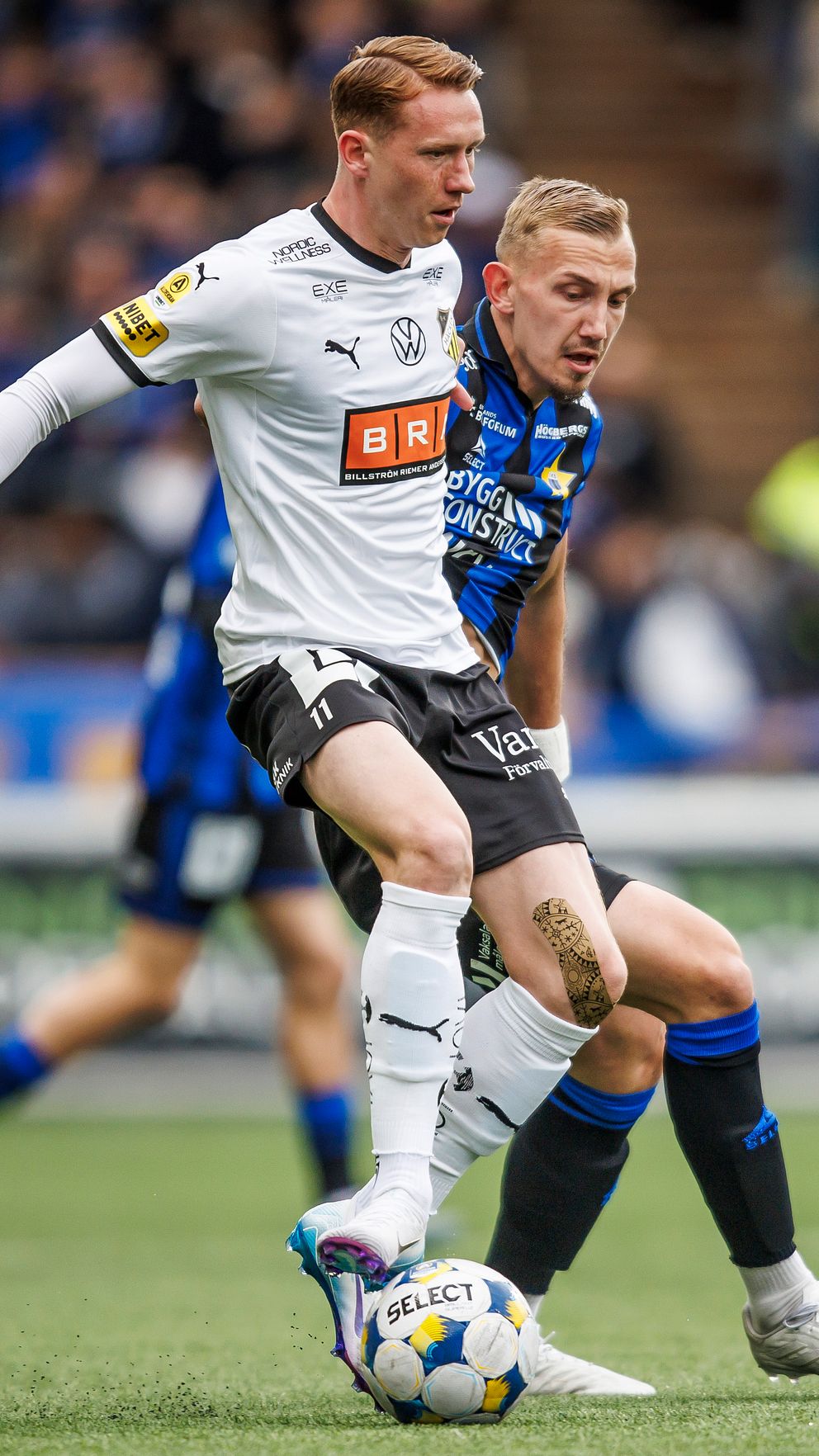 Två fotbollsspelare kämpar om bollen under en match på gräsplan med publik i bakgrunden.