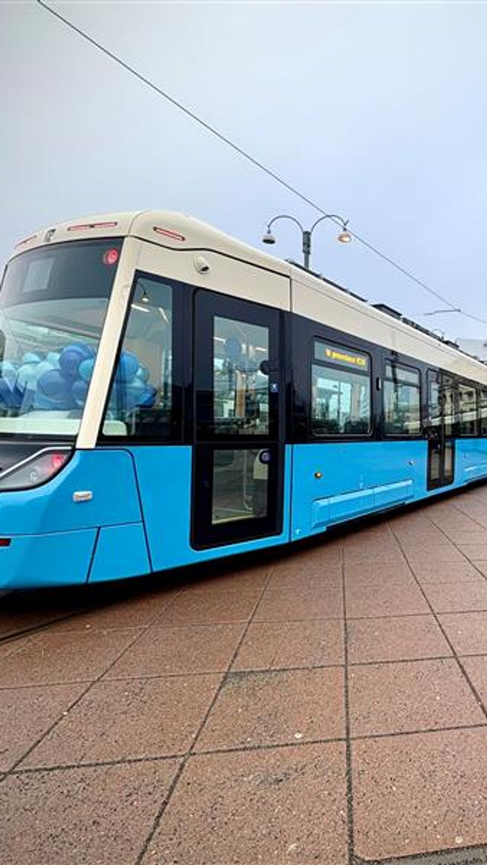 En modern spårvagn i ljusblå färg står på en torgyta med stora plattor. I bakgrunden syns historisk arkitektur och några personer som går förbi, med en lätt dimma över området.