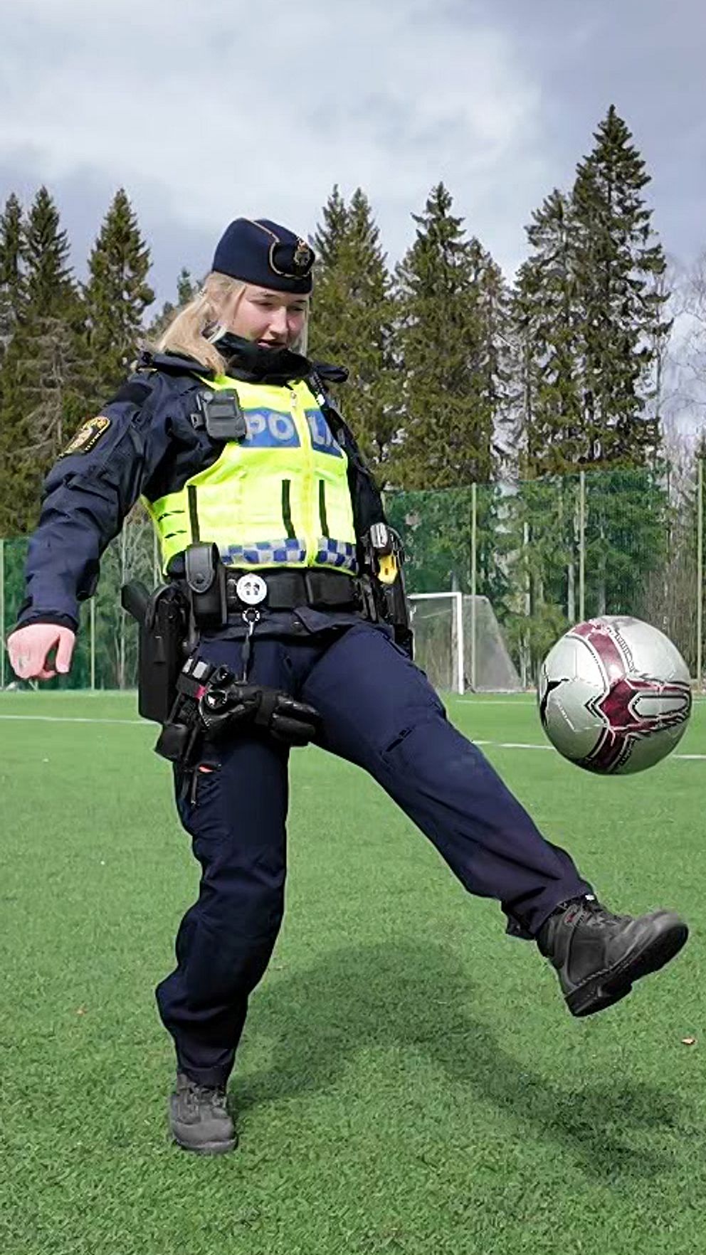 En polis jonglerar med en fotboll på en gräsplan utomhus framför träd och skog.