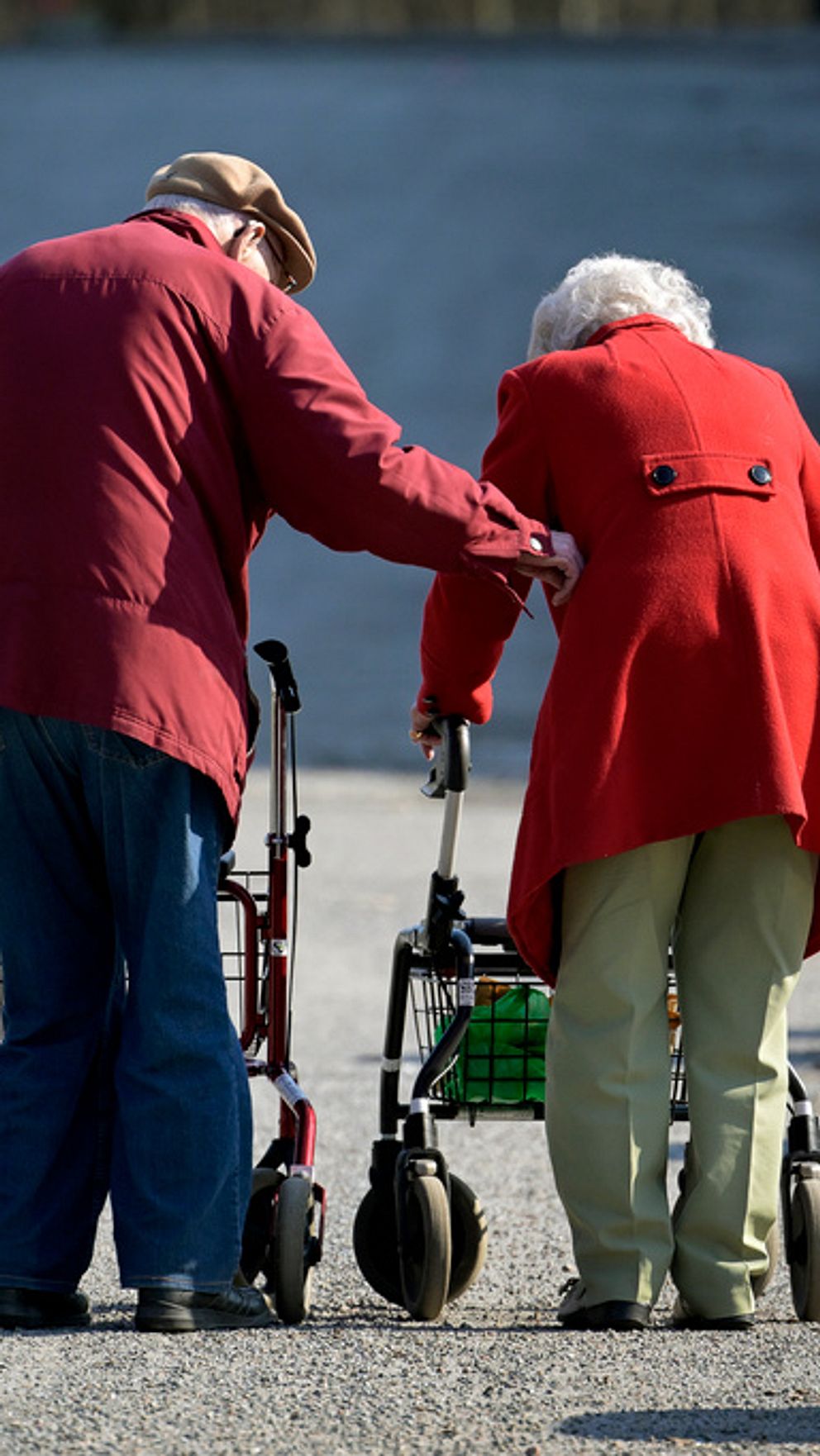 Pensionärspar klädda i rött med varsin rollator. Mannen håller en stöttande hand på kvinnan.