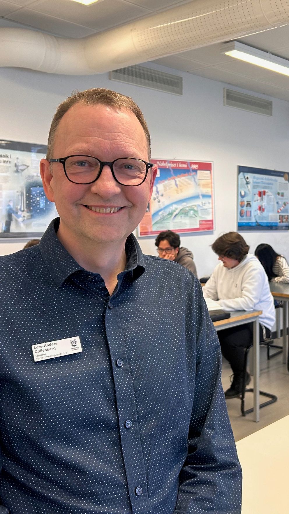 Klassrum med elever vid skrivbord och en lärare i förgrunden. han ler mot kameran.