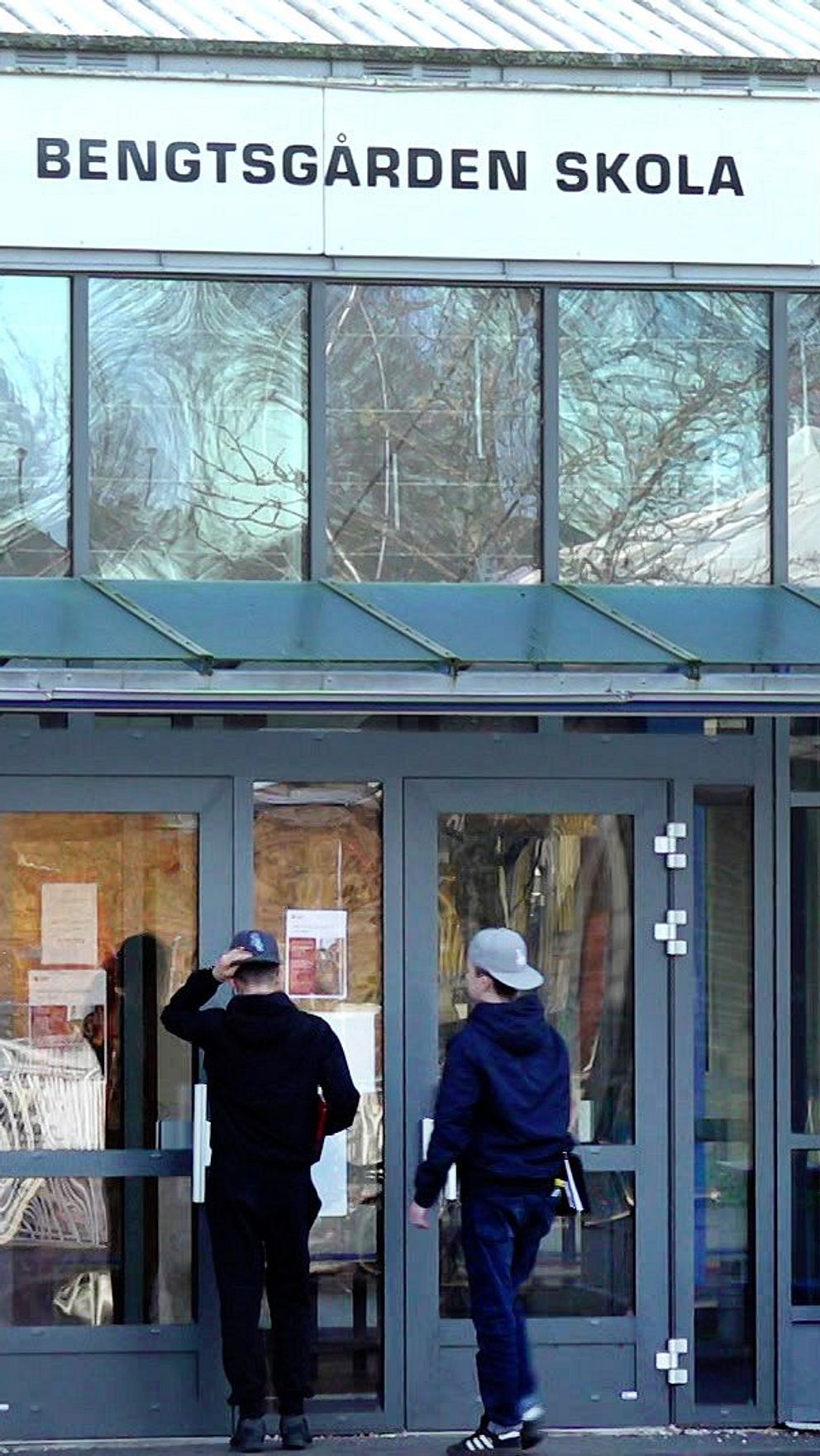 Entrén till Bengtsgården Skola, med stora fönster och en glasad dörr. Två personer står vid ingången. På en n skylt ovanför dörren står skolans namn.
