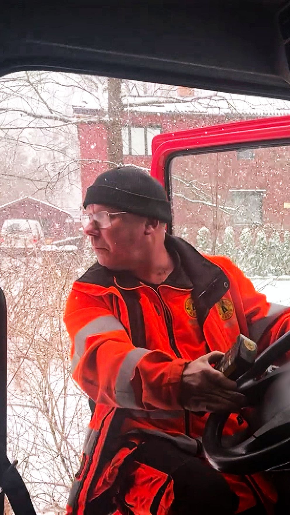 En bärgare iklädd en orange jacka hoppar ur en bärgningsbil. Bilens instrumentbräda och kontroller är synliga inuti hytten, och utanför snöar det.