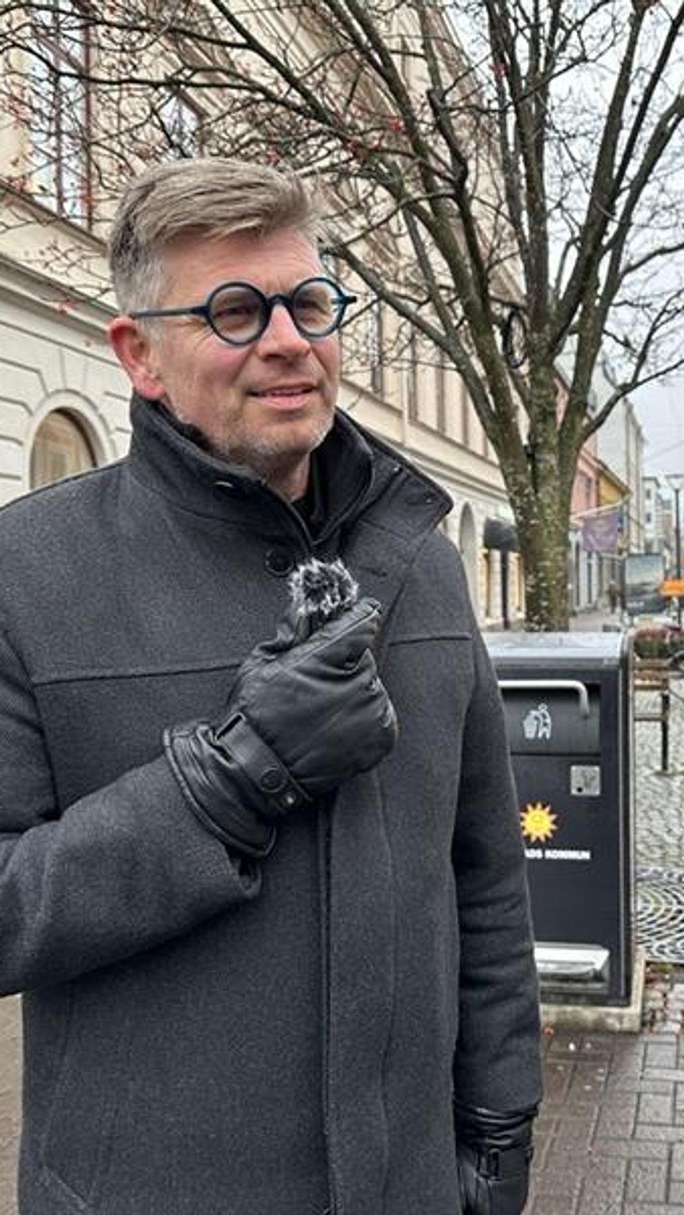 Malte Hallquist står på en gata i Karlstad och håller upp en mick i handen. Framför personen finns en smartphone monterad på ett stativ, som visar en skärm med en spegelbild av personen. Bakgrunden består av byggnader och träd, vilket ger en stadsatmosfär.