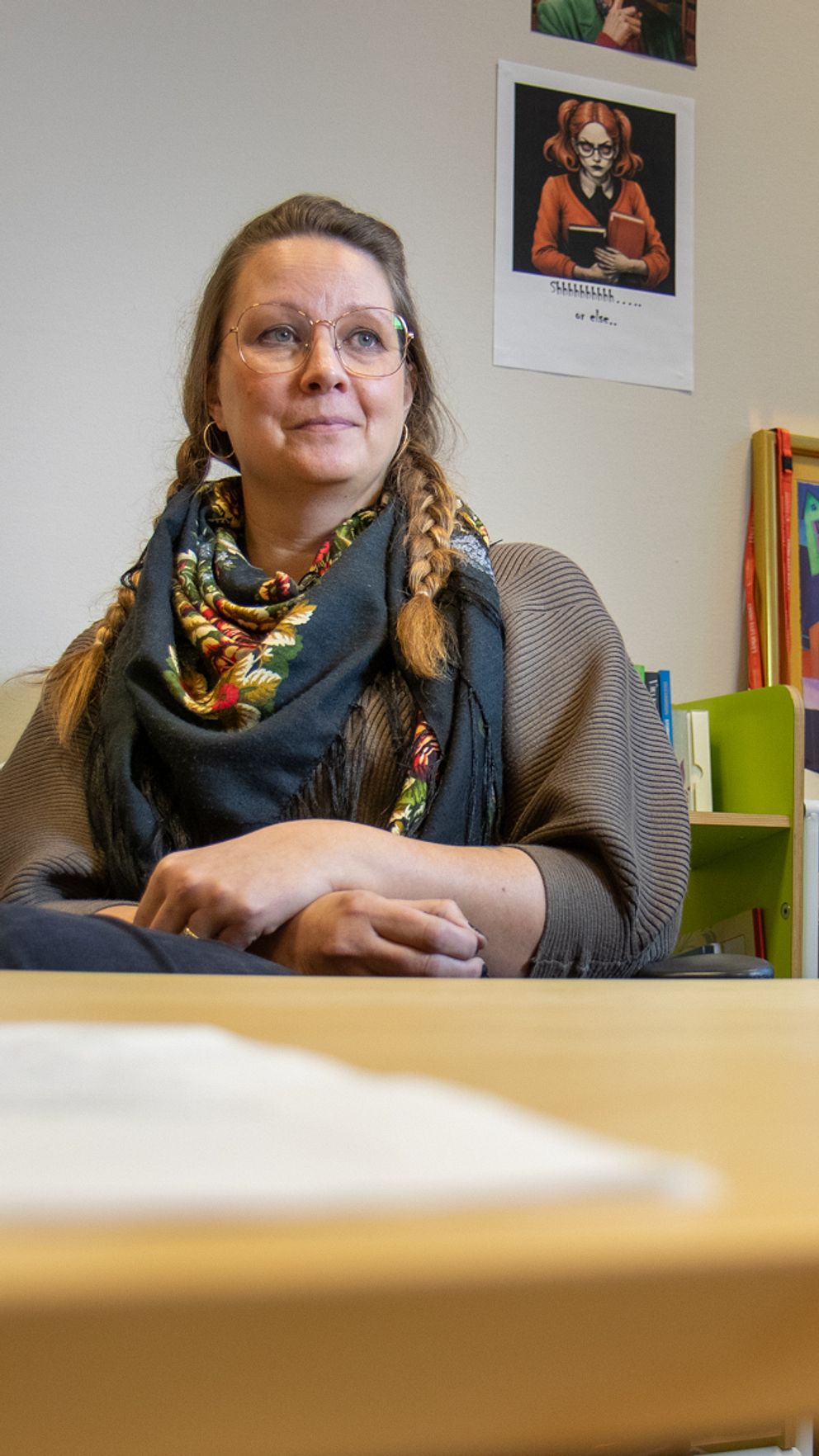 En person sitter vid ett träbord i en biblioteksliknande miljö, omgiven av böcker och konstverk på väggarna. Framför personen ligger ett papper och en informationsskylt. Rummet är ljus och inbjudande, med element av färgglad konst och en grön växt i bakgrunden.