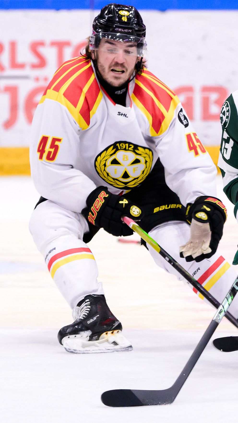 Två ishockeyspelare i matchdräkter från olika lag kämpar om pucken på isen, en i vita och röda färger med nummer 45 och den andra i gröna med nummer 15. Bakgrunden visar en del av ishockeyrinken med reklam.