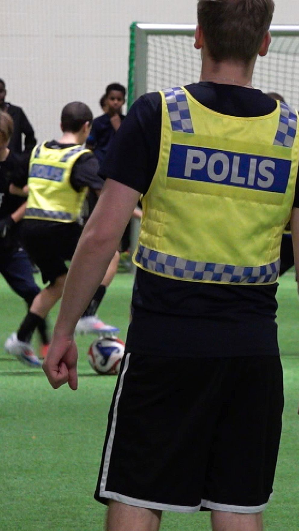 En grupp ungdomar spelar fotboll inomhus, medan en polis, klädd i en reflexväst med texten ”POLIS”, övervakar aktiviteten. I bakgrunden syns fotbollsmål och fler spelare som deltar i matchen.