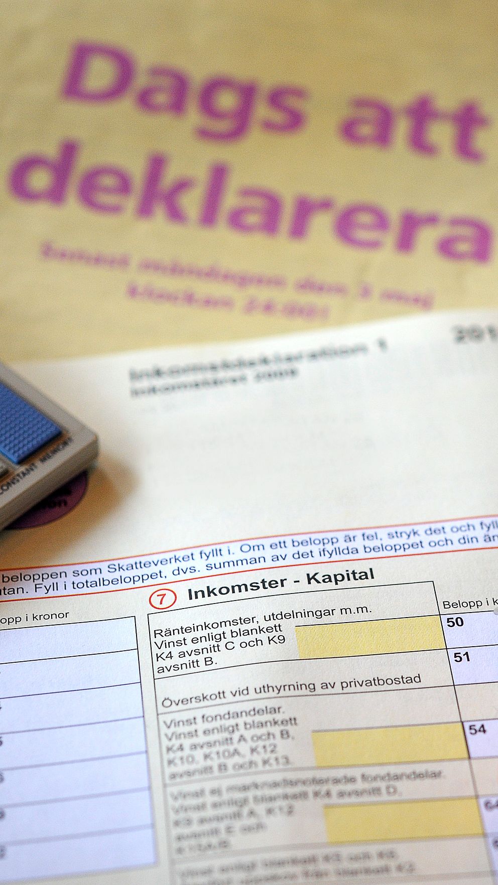 Person fyller i en deklarationsblankett för inkomst av kapital med en penna. I bakgrunden ligger ett informationsblad med texten ”Dags att deklarera” samt en miniräknare, vilket indikerar att det handlar om att lämna in årlig deklaration.