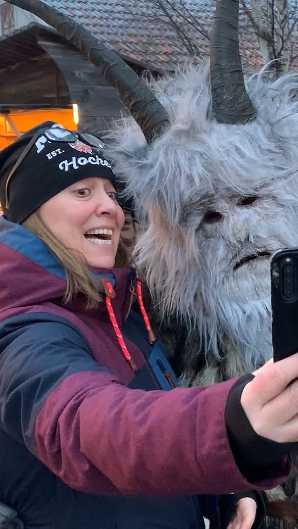En person står nära en kostymklädd figur som ser ut som ett mytologiskt väsen med horn och en pälsig dräkt. De tar en selfie tillsammans i en livlig, vinterlig miljö. I bakgrunden syns flera besökare och en byggnad dekorerad med ljus. Stämningen är festlig och social.