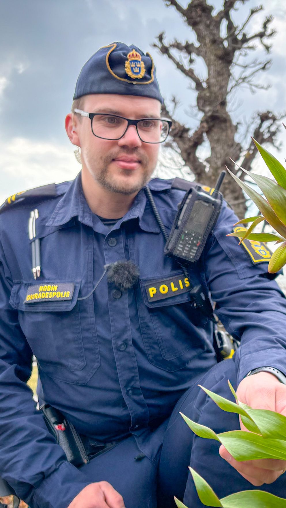 En polisinspektör knäböjer vid växt utomhus, med träd och himmel i bakgrunden.