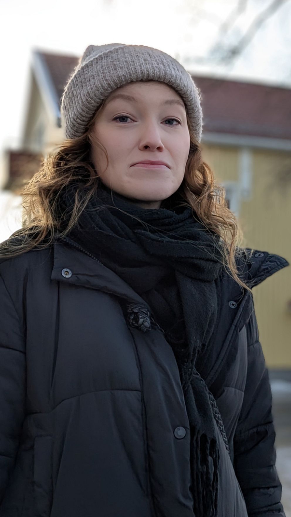 Mia står på en snötäckt gata i Skellefteå med en gul byggnad i bakgrunden, omgiven av träd. Det är en klar dag med solsken. Mia bär en svart jacka och en ljus mössa, med halsduk som skyddar mot kylan.