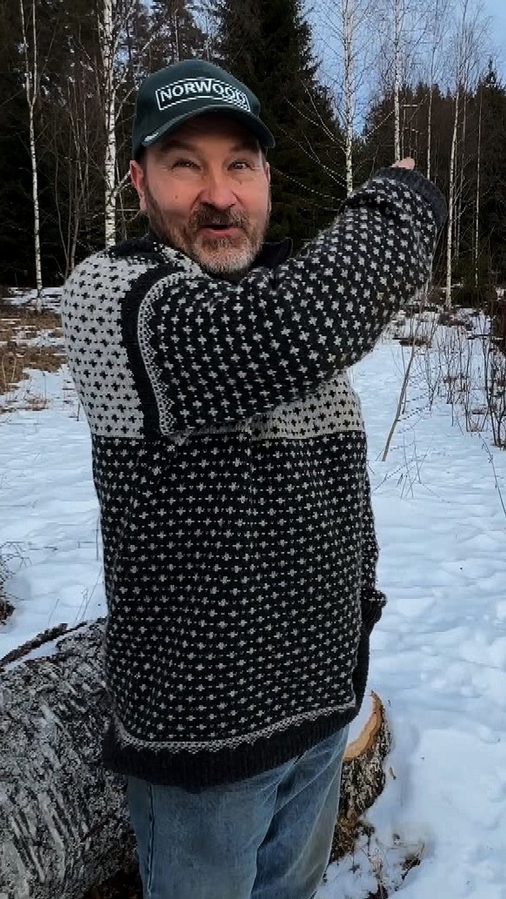 En person i stickad tröja pekar mot en snötäckt trädgård vid en stuga.