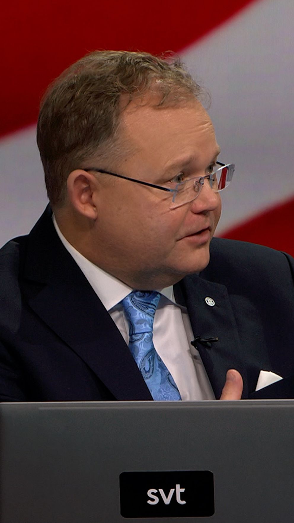 En man i kostym gestikulerar medan han pratar vid ett podium med två bärbara datorer framför sig.