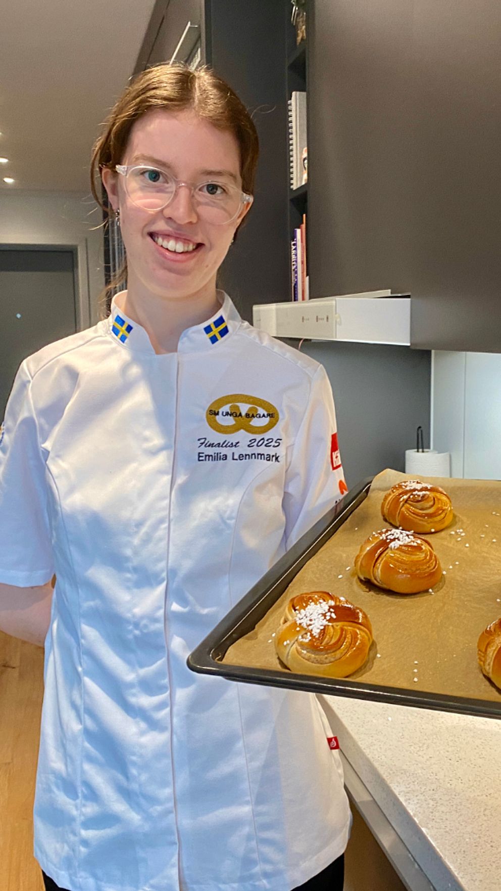 Konditor visar en plåt med nybakta kanelbullar i modernt kök.